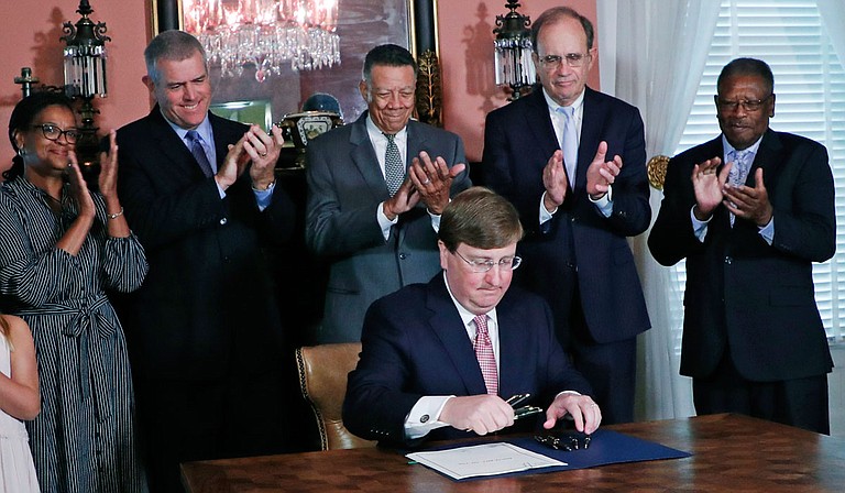 Gov. Tate Reeves signs the bill to remove the state flag. House Speaker Philip Gunn and Lt. Gov. Delbert Hosemann, both now infected with COVID-19, stand behind him. Dr. Thomas Dobbs now says Mississippi needs mask mandates. Photo by Rogelio V. Solis via AP